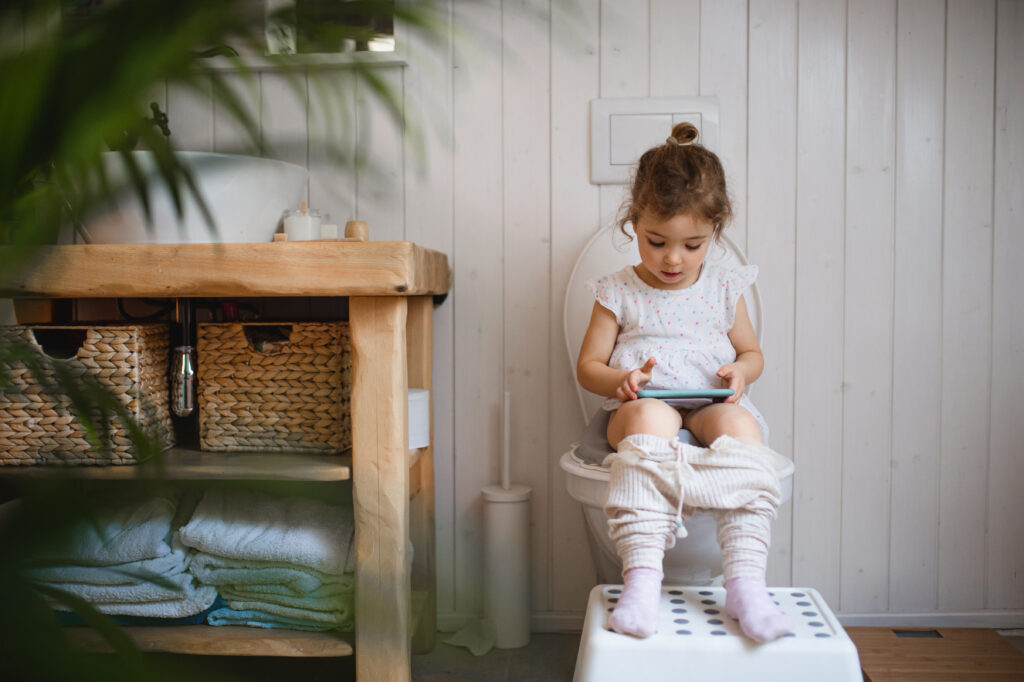 Portrait of cute small sitting on toilet indoors at home, using smartphone. little girl sitting on the toilet and reading