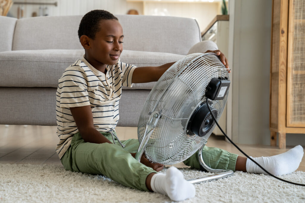 Calm African boy refreshing with electric fan at home alone. Small boy enjoy strong fresh wind blow child sitting infront of fan to cool down