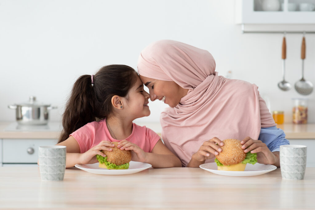 Lunch With Mommy. Little Girl Eating Sandwiches With Muslim Mother In Kitchen mother and child happy with a meal