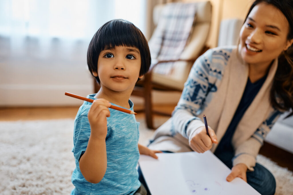 Small Asian boy coloring on the paper with his mother at home.