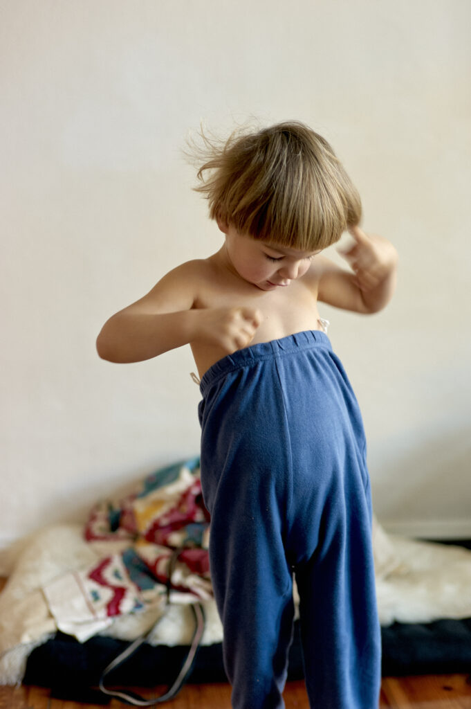 Child trying to dress themselves standing in a room with neutral background. child trying to put on his pants