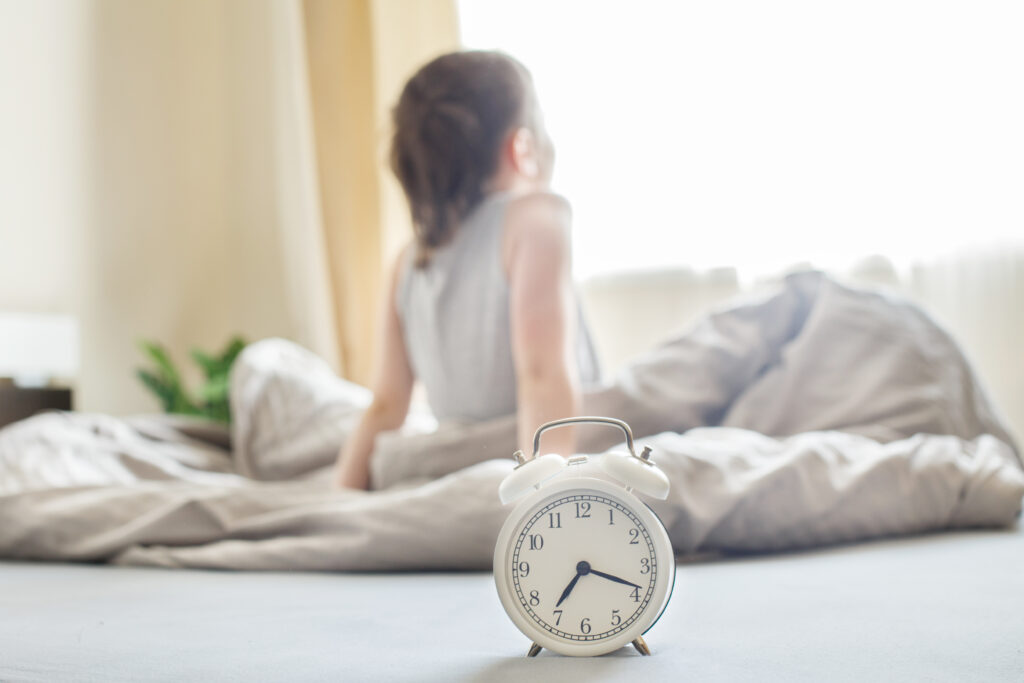 little boy sitting in bed at home in the morning on a window background with alarm clock